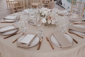 The round tables with simple, neutral linens, linen napkins, and a beautiful, low, elongated arrangement with roses, spray roses, ranunculus, scabiosa, baby's breath and bleached ruscus which had been used down the aisle in the ceremony.
