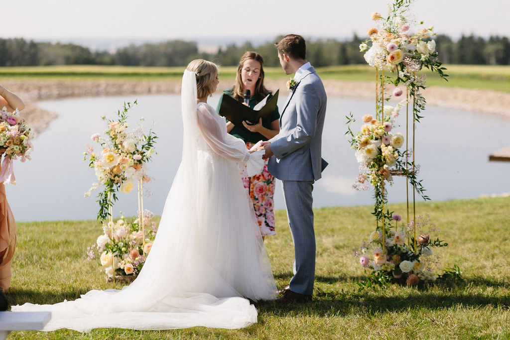 McKinley and Eric with clasped hands during the wedding ceremony, with the floral stands on either side.