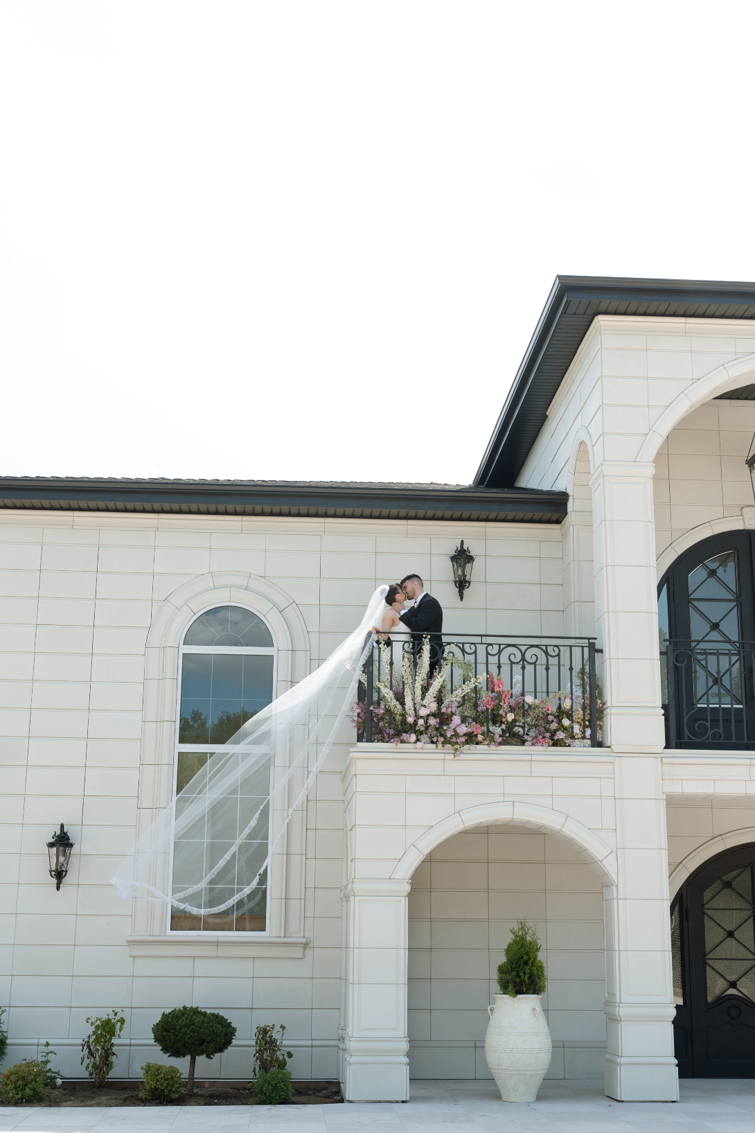 Couple stands on the balcony at Archway Manor with her long veil cascading over the railing.