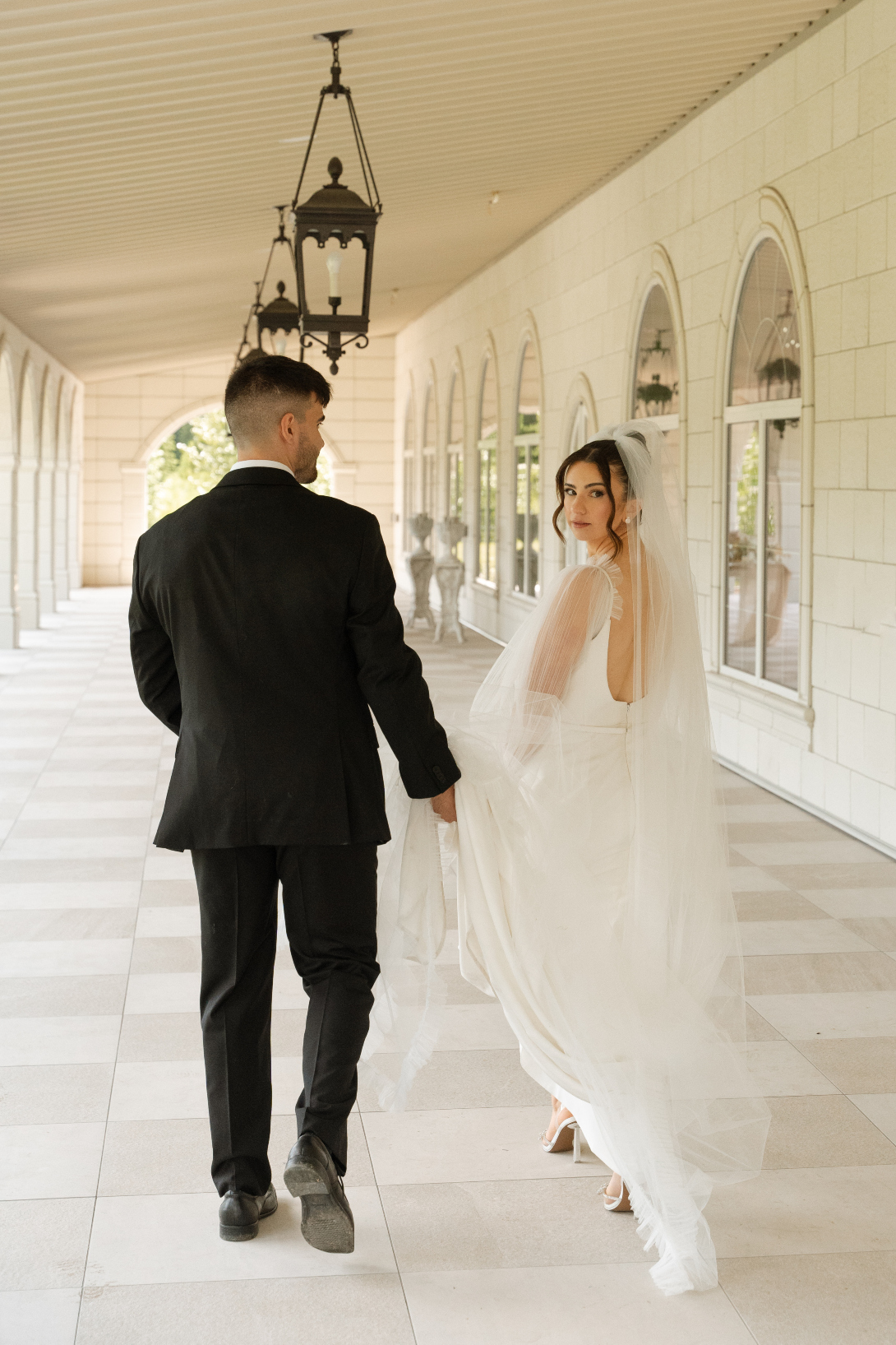 The couple strolling along the outdoor terrace, with many archway pillars to the left with views of the pond.