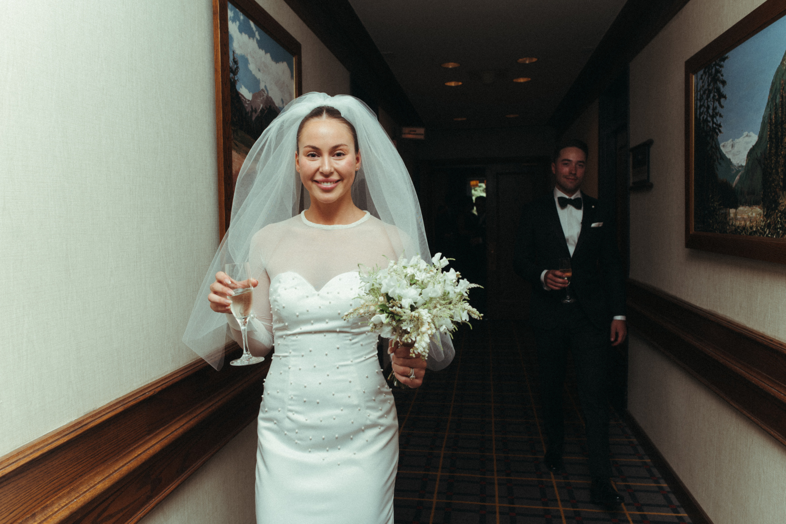 Tyller walking down the hallway with a large smile, a glass of champagne in one hand and her bouquet in the other, while Carter is following behind her in the background.