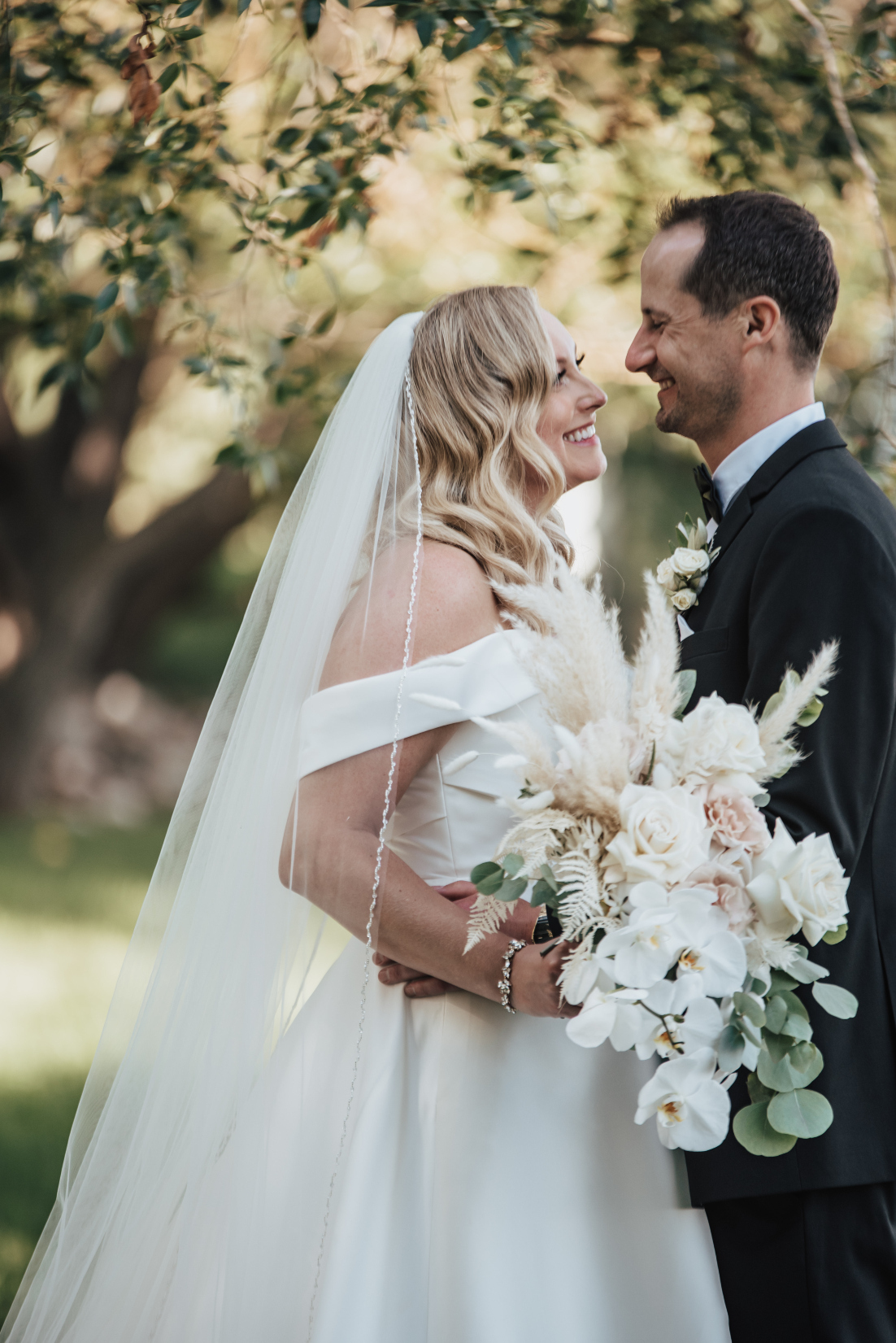 Holly and Scott are standing closely, gazing at each other and smiling. Holly holds her beautiful bouquet at her side.