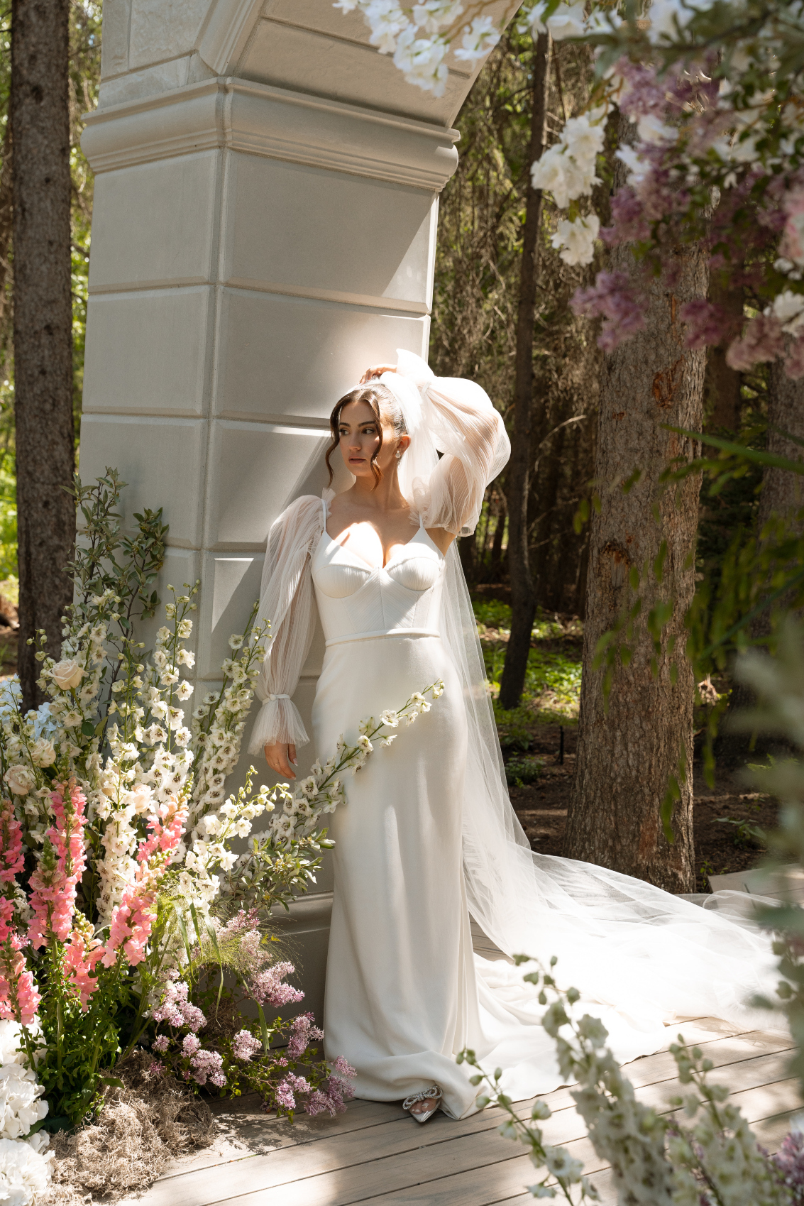 The bride leans against the stone archway with one hand above her head, the other at her side as the sun shines on her and the flowers at her feet.