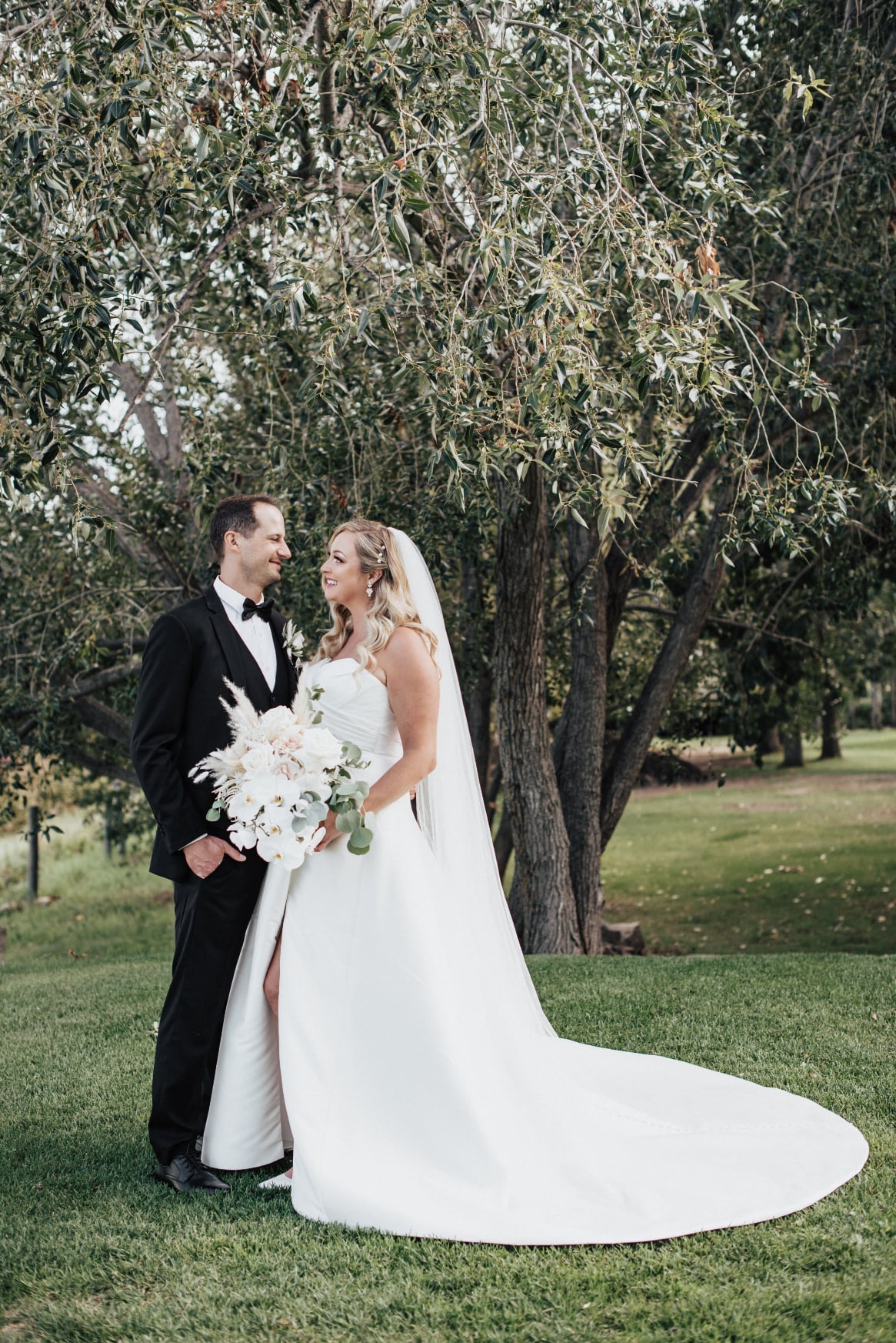 Holly and Scott are standing closely in front of a lovely tree, looking lovingly into each others eyes.