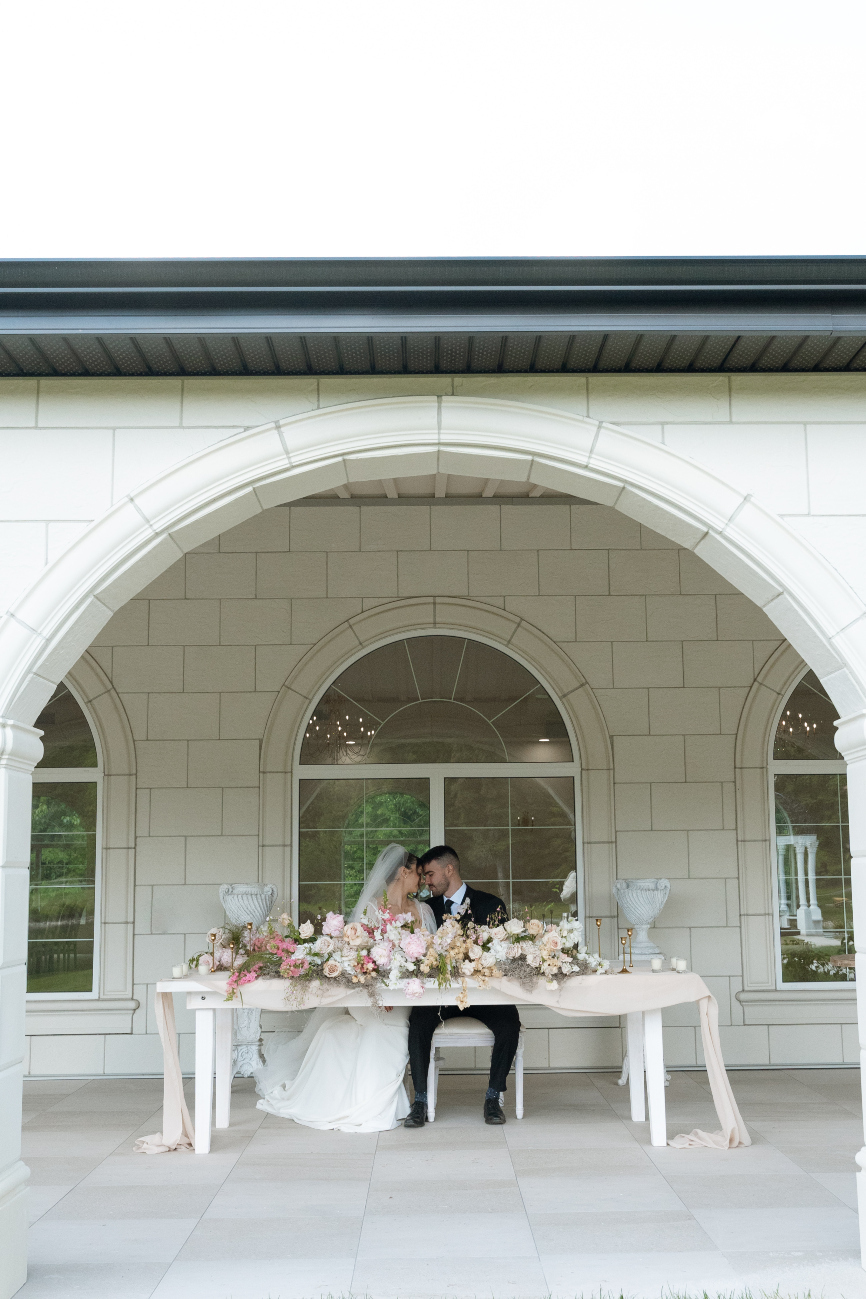 A view looking through one of the arches into the terrace where the couple sits with their heads tipped together at a sweetheart table adorned with a long and low floral arrangement.