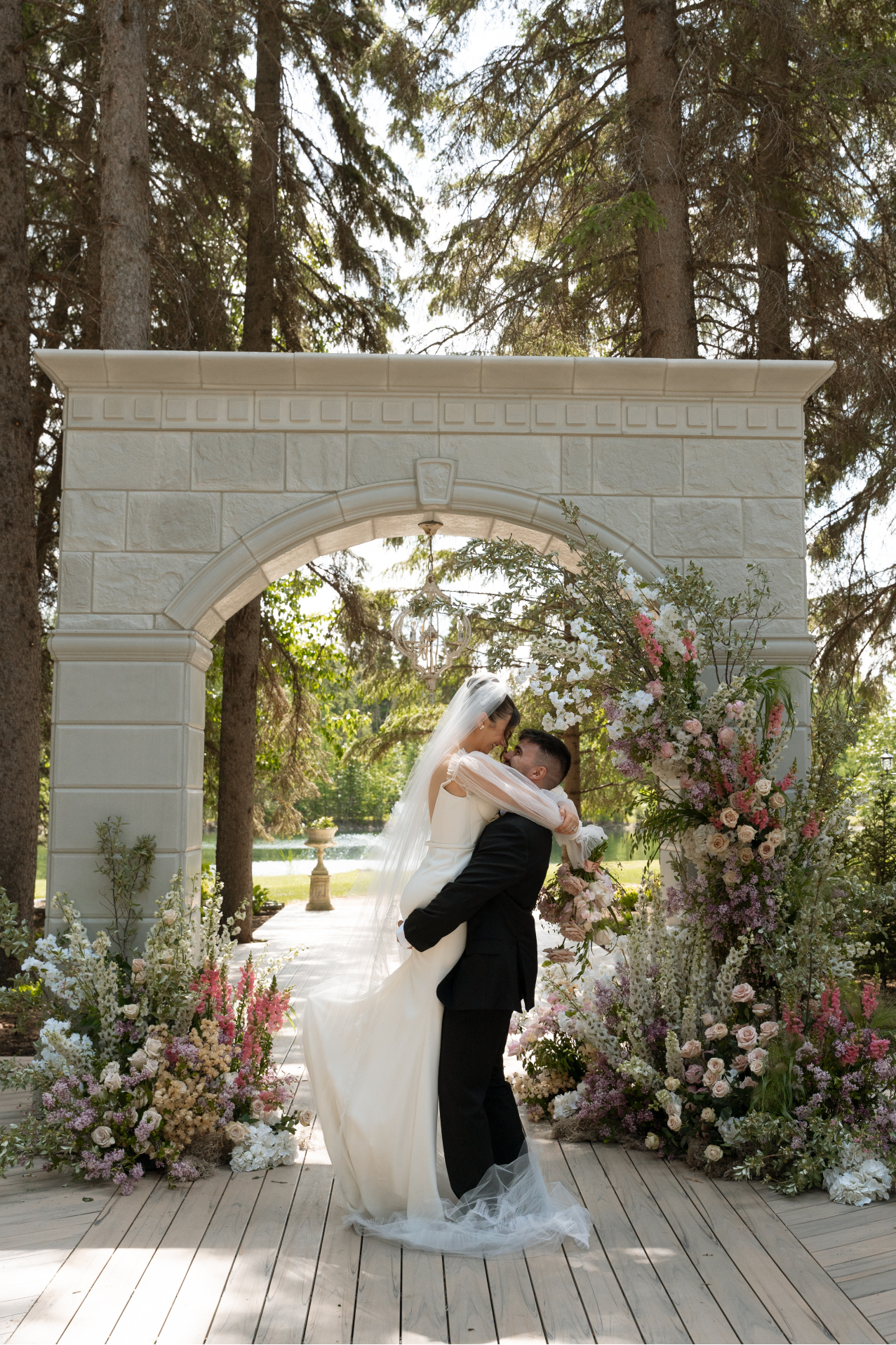 The groom is lifting the bride off her feet as they embrace under the stunning stone archway at the ceremony site, which is adorned with gorgeous flowers.