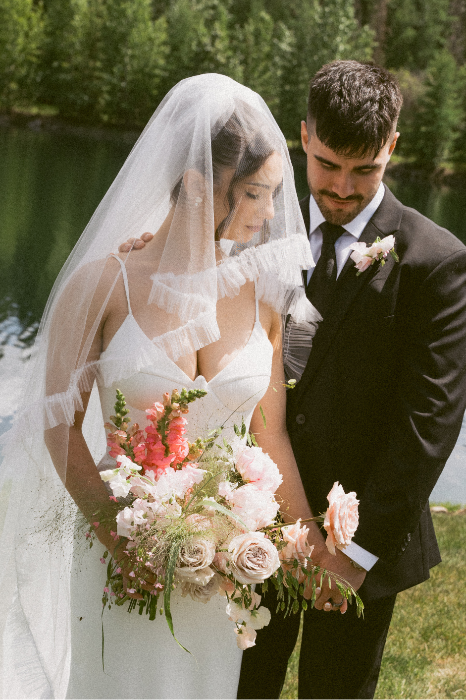 The bride and groom are holding hands and are snuggled up closely to one another in front of the pond