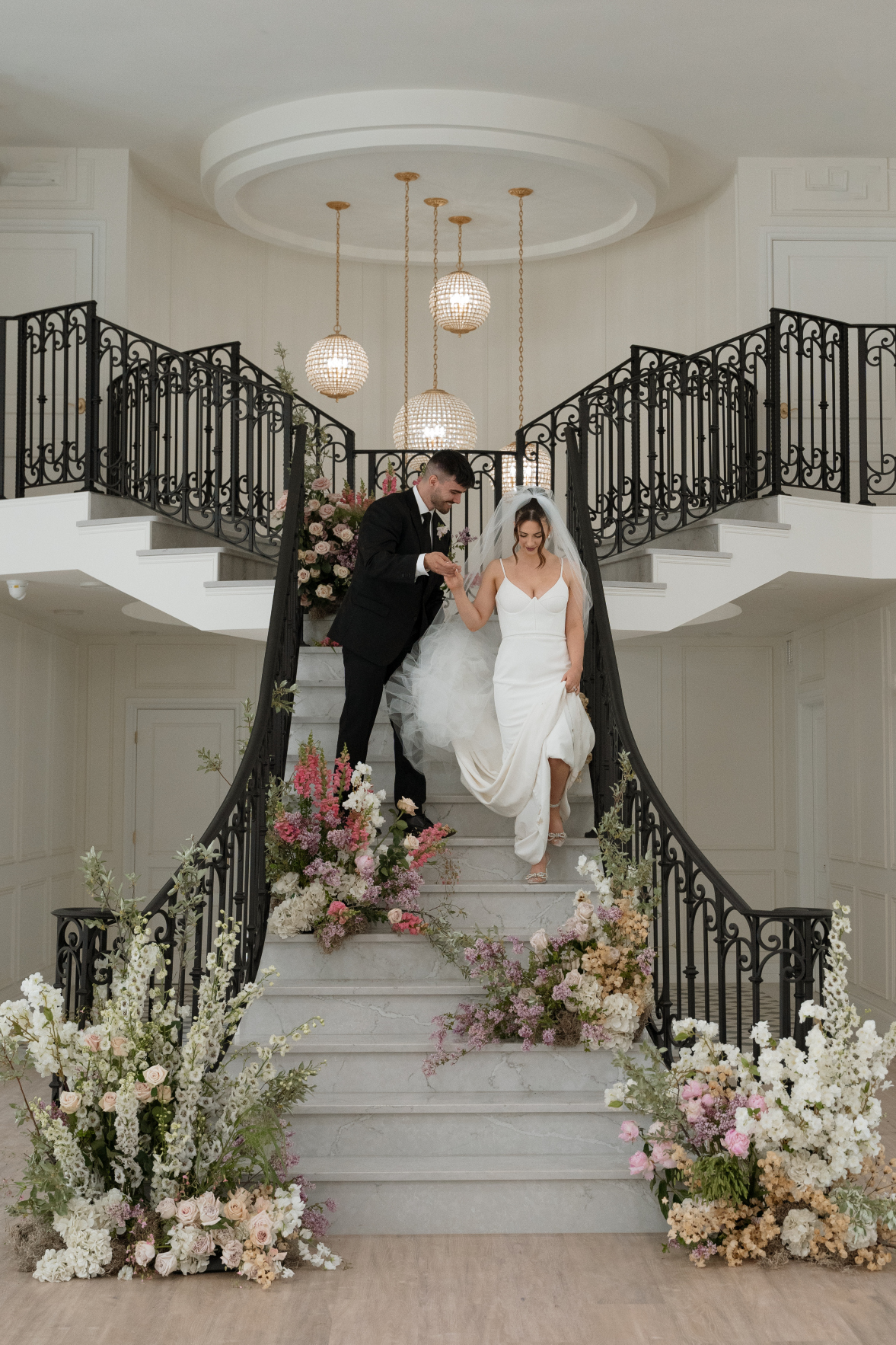 The groom holds the brides hand, helping her down the grand staircase, which is adorned with several floral arrangements