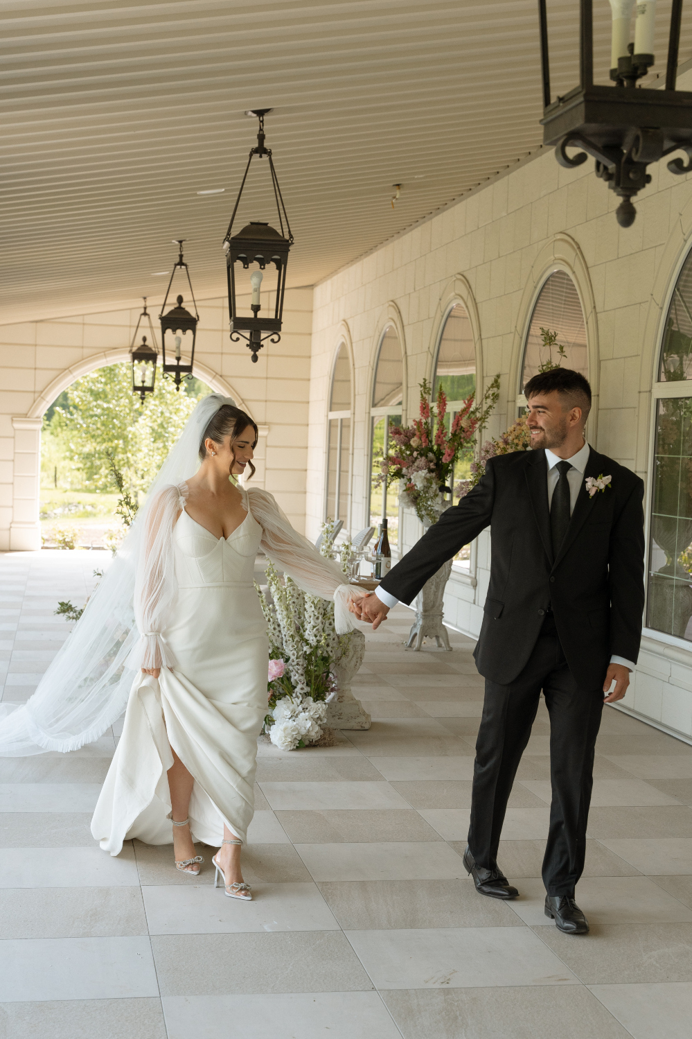 The couple walking hand in hand out on the terrace, with beautiful tile floors, arched windows, and hanging light fixtures.