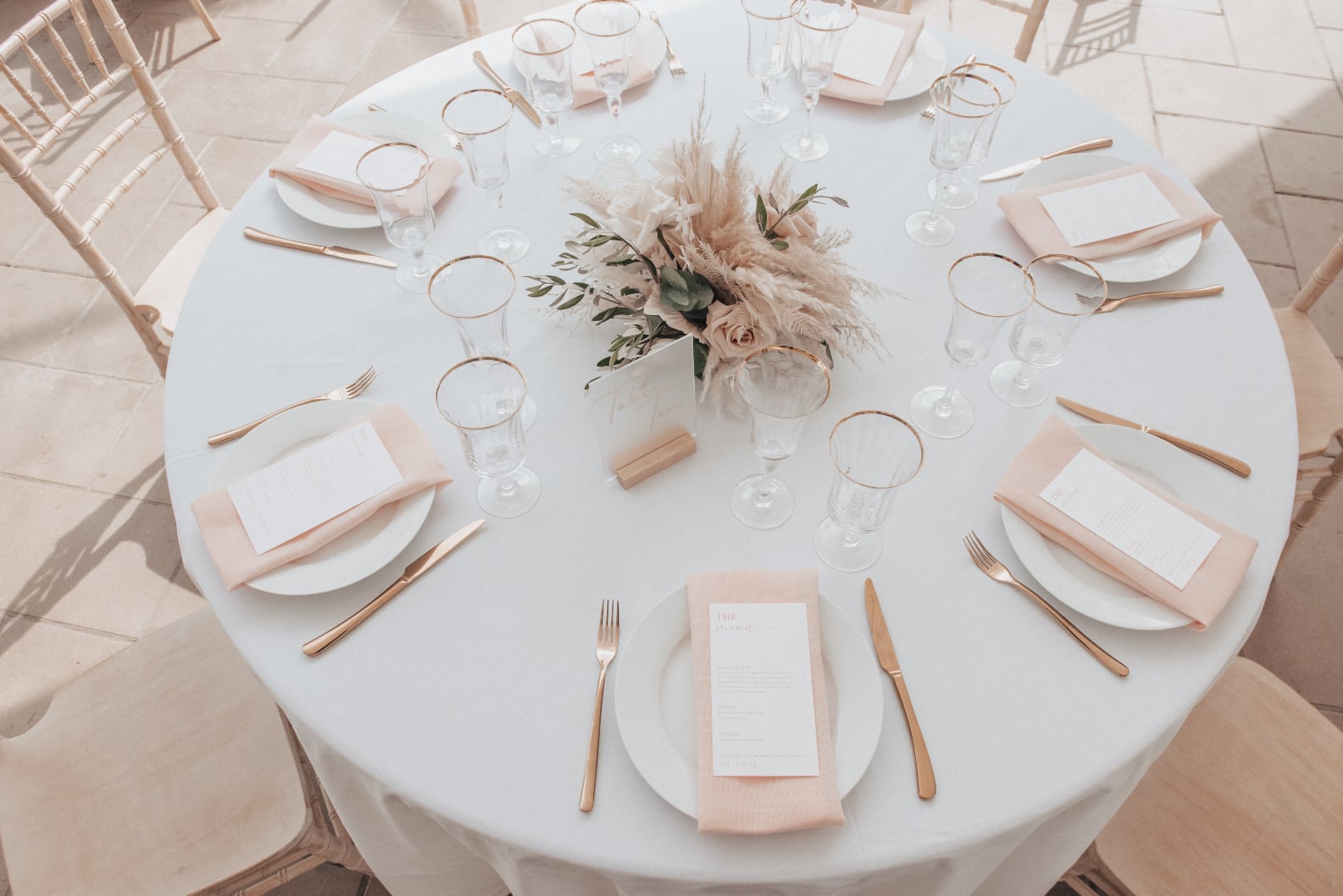 A high angle shot of the guest tables, showing the simple white linens, blush napkins, floral centrepiece and golden accents of the glassware and flatware.