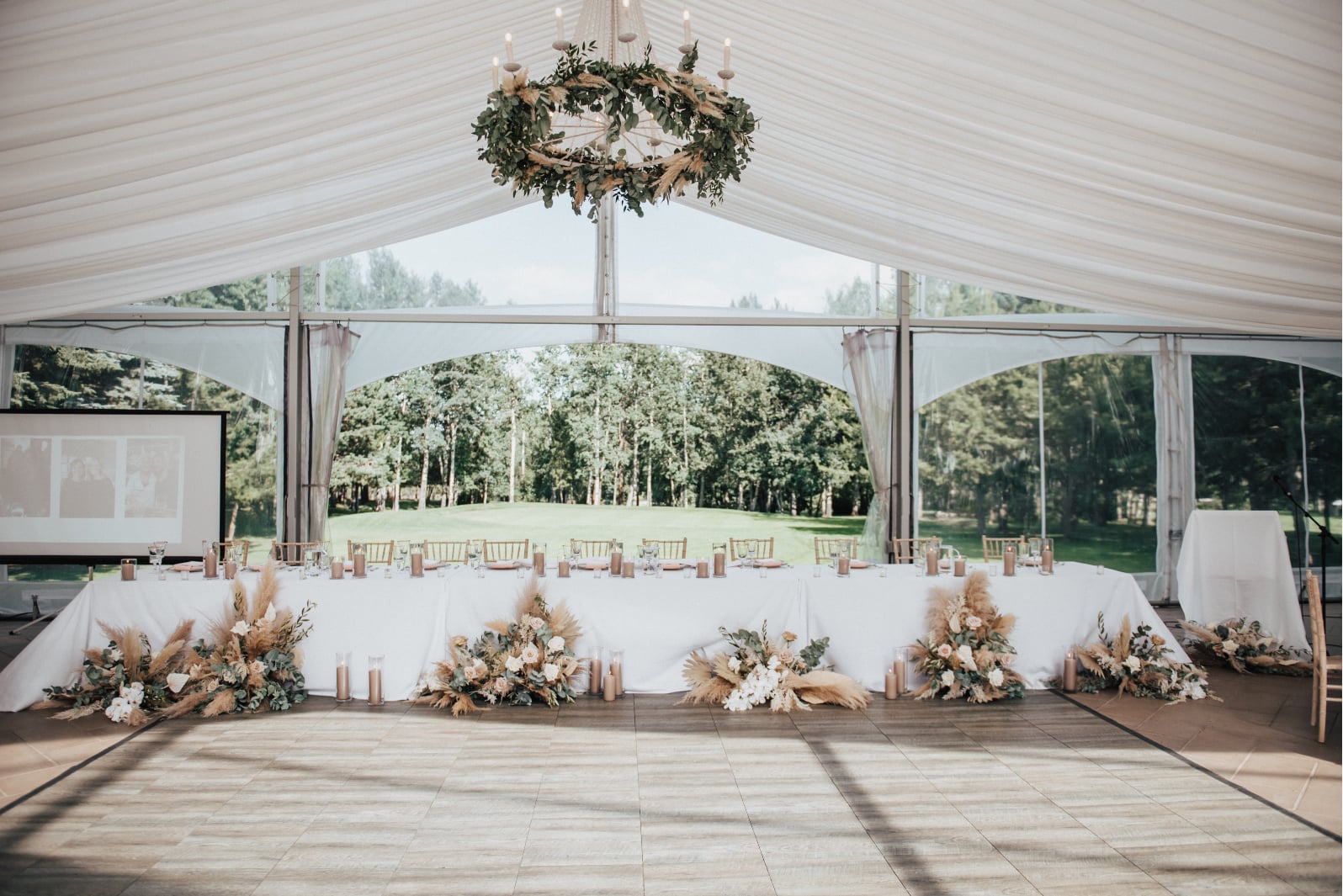 The view of the head table, with greenery chandelier, tan pillar candles running down the length of the table at varying heights and large floral arrangements and pillar candle clusters on the floor in front of the table.