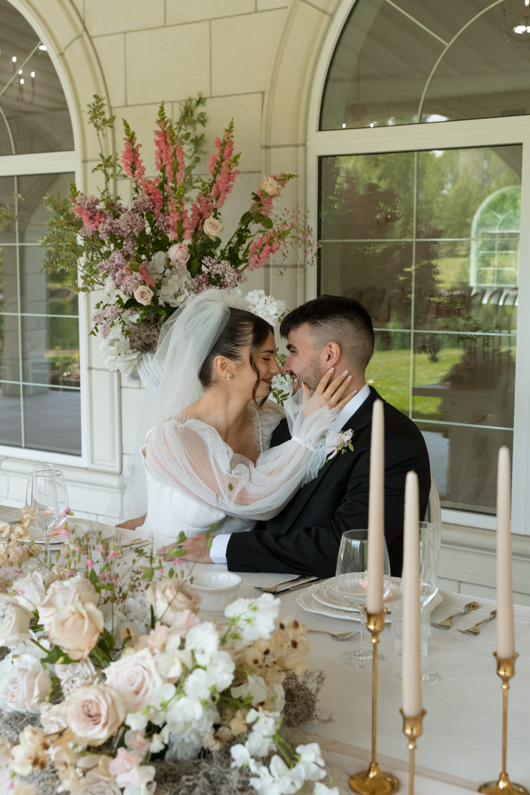 The bride and groom are sitting at the sweetheart table facing one another, the bride is cupping the grooms face with her hands, with a large floral arrangement in the background.