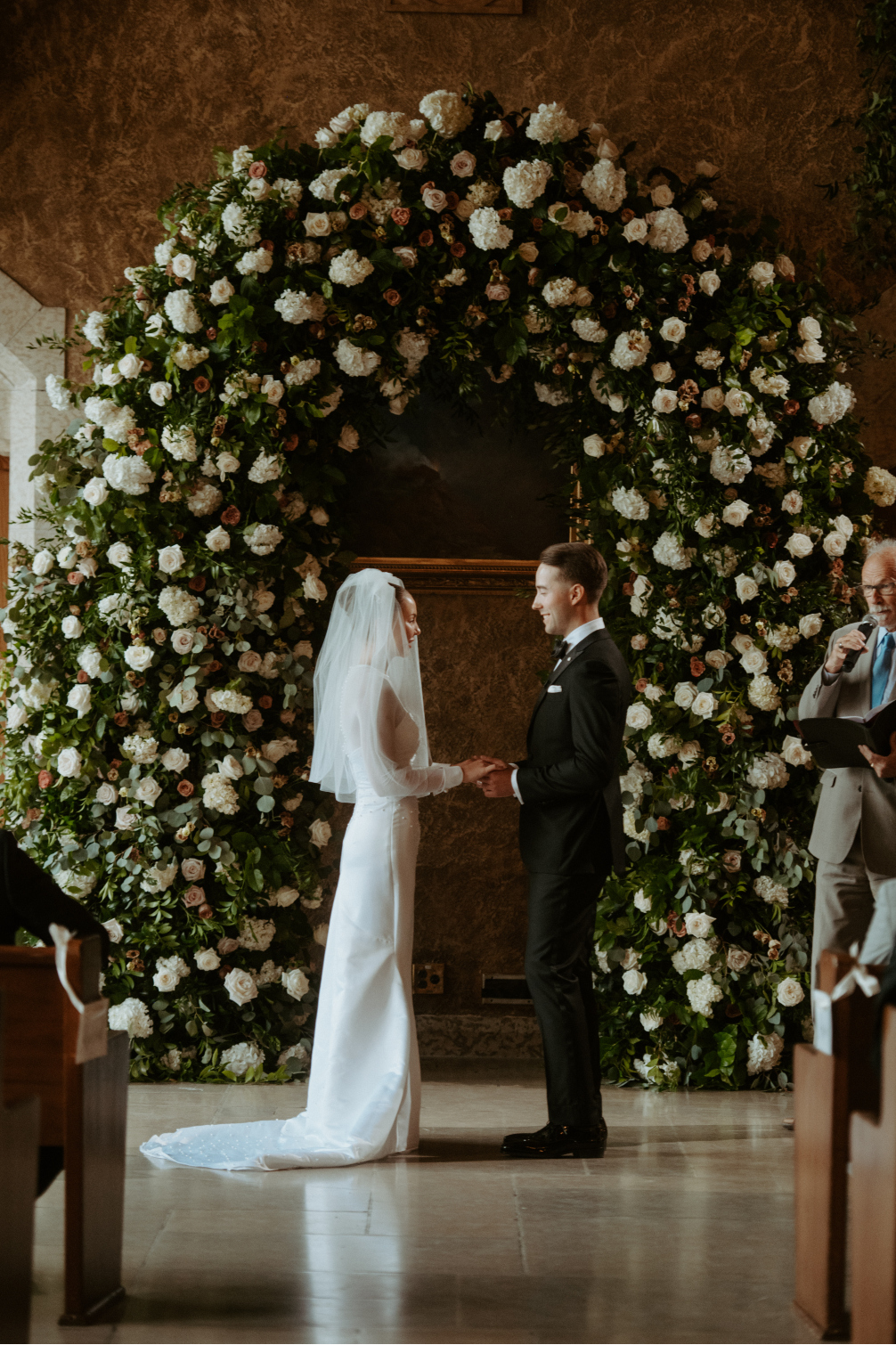 Tyller and Carter face each other with clasped hands during the ceremony in front of the big beautiful floral arch.