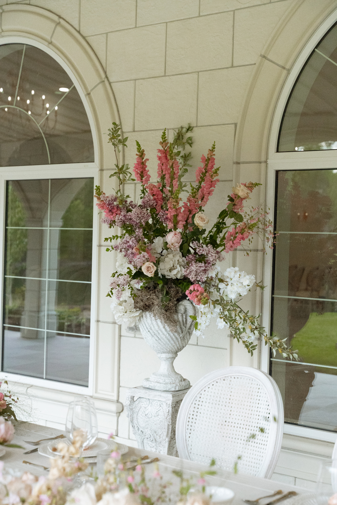 A photo of one of the arrangements in a pedestal vase on top of a pillar, including pink snapdragons, hydrangea, lilacs, pink peonies, roses and cherry blossom.