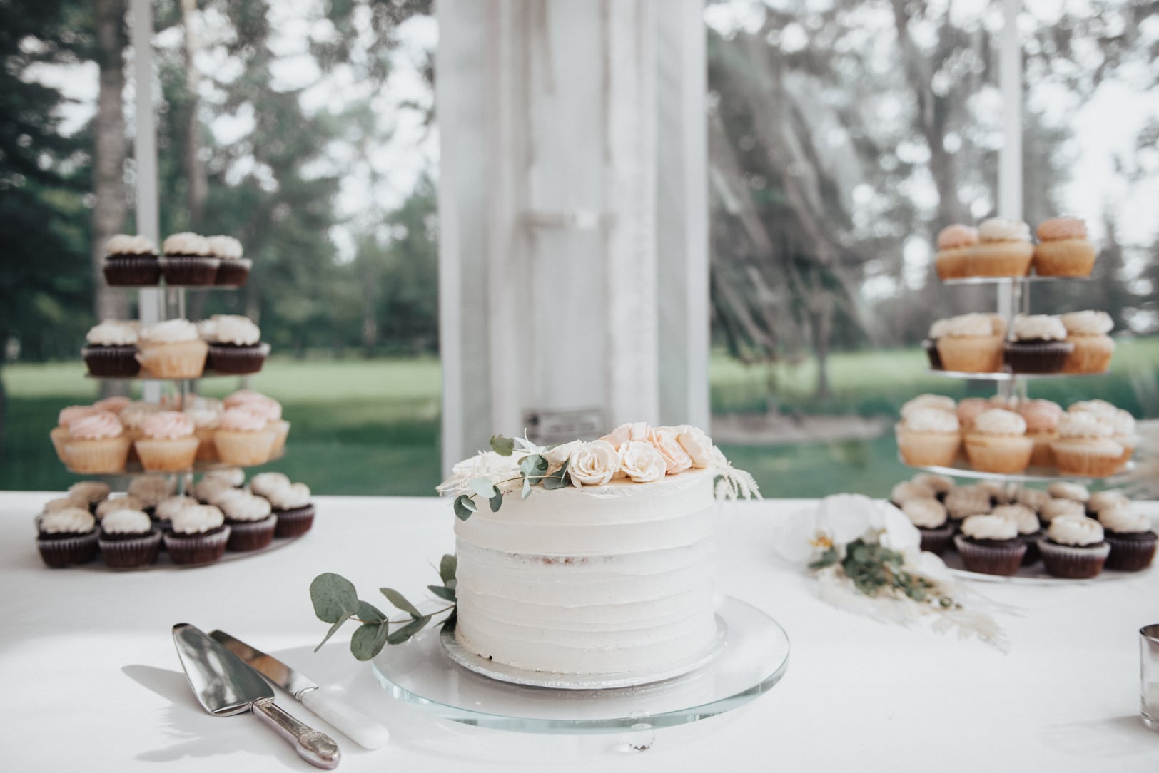 A simple, single tier cake with buttercream flowers and eucalyptus with towers of cupcakes behind.