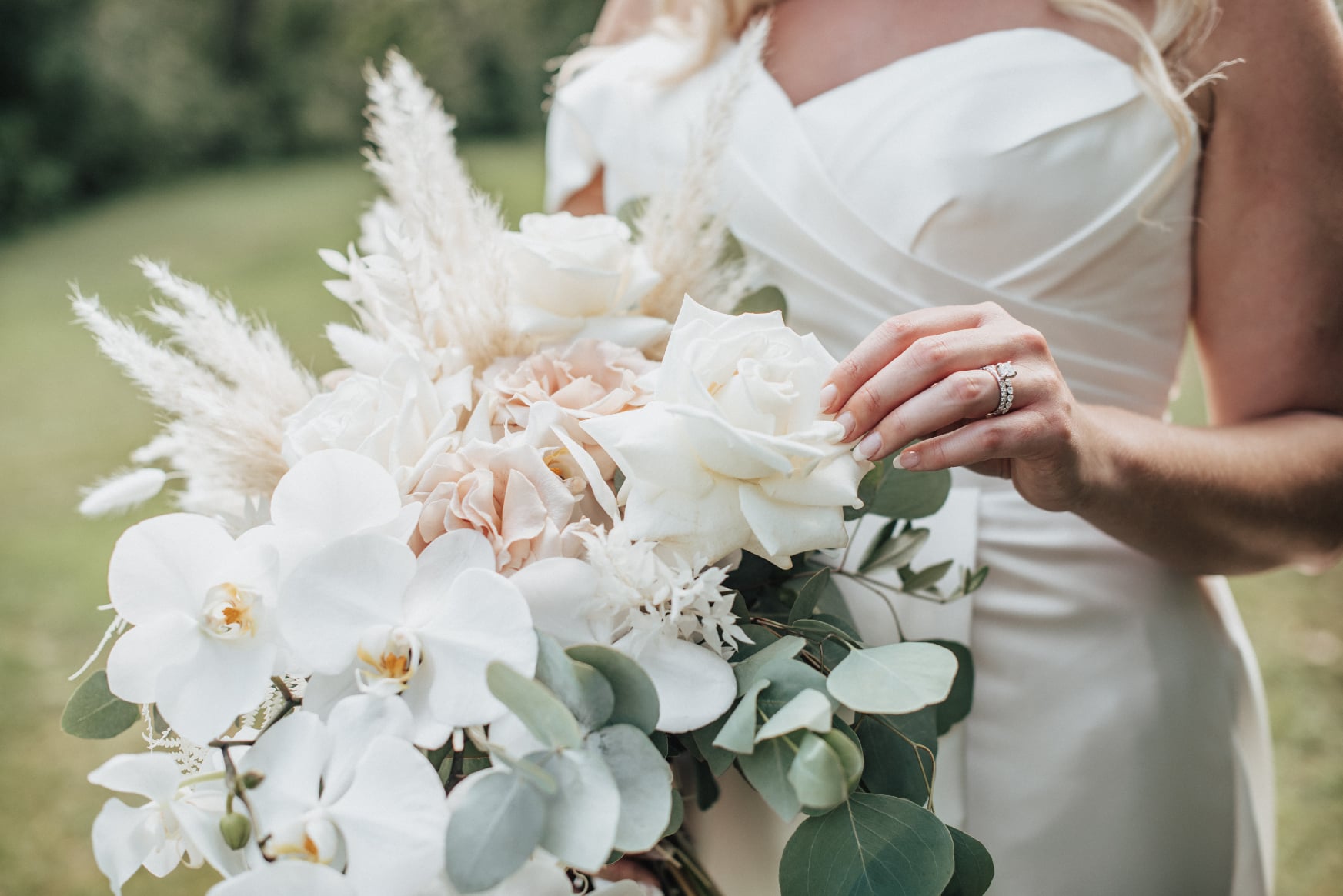 A close up image of the bridal bouquet with gorgeous white orchids, full roses, pampas grass, bleached bracken fern, olive branch and eucalyptus. Holly is gently feeling the soft petals of one of the roses.