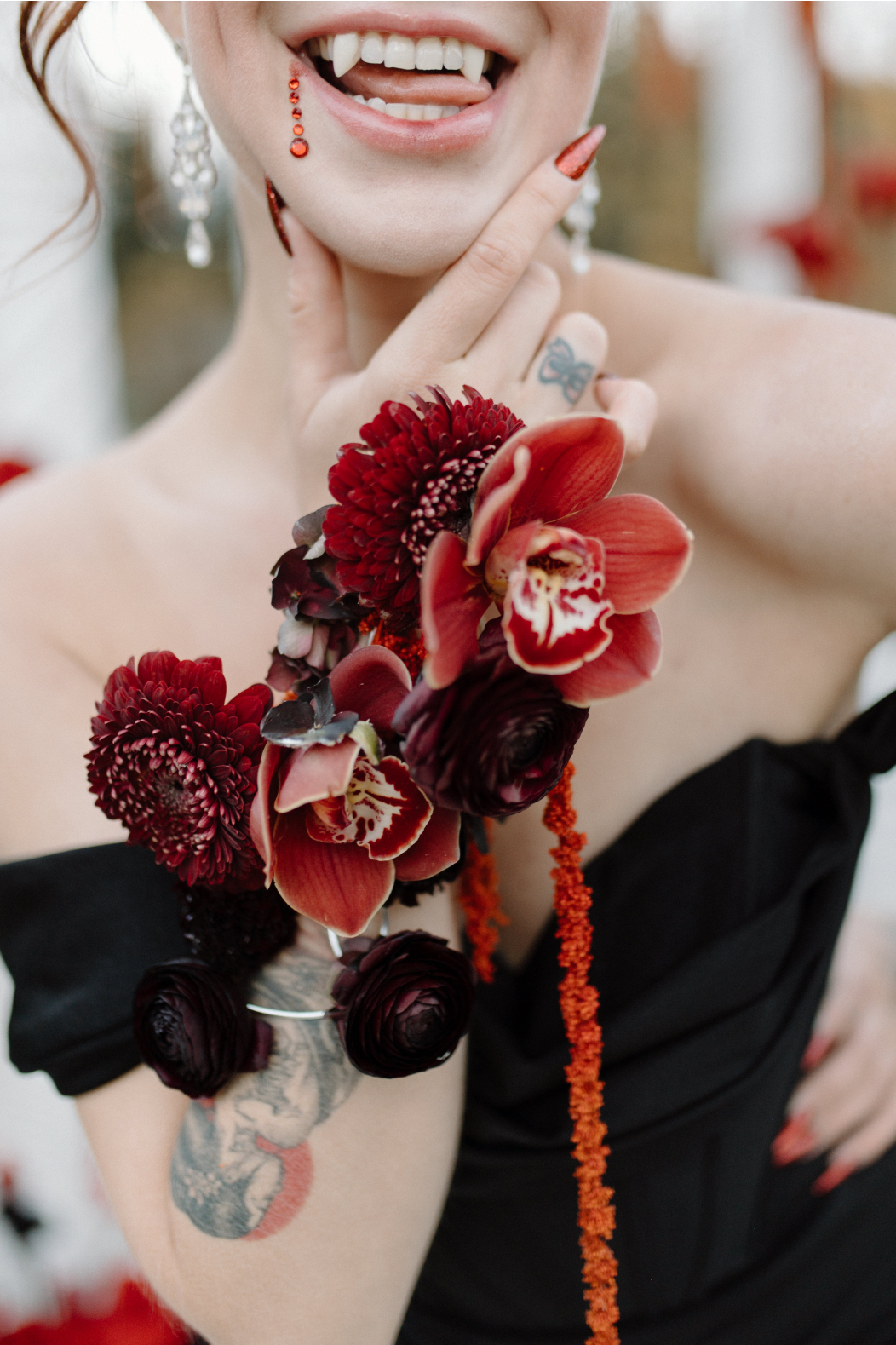 Dracula's bride, holds her hand up to her chin, showing off a stunning, elaborate wrist corsage designed in reds and black. She sticks her tongue out of the side of her mouth in a cheeky expression, showing off her fangs.