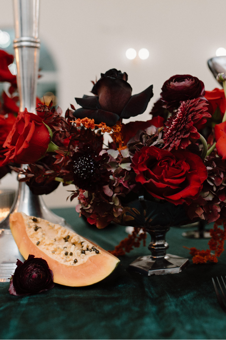 Close up of a beautiful centrepiece in a pedestal container featuring red roses, burgundy ranunculus, black scabiosa, red amaranthus, red gerbera, and a sliced open papaya sitting on the table beside it.
