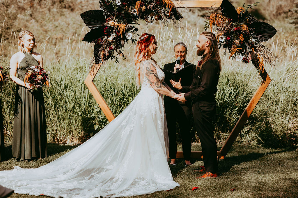 Haley and Greg during their ceremony, clasping hands in front of the beautiful hexagon arch adorned with flowers.
