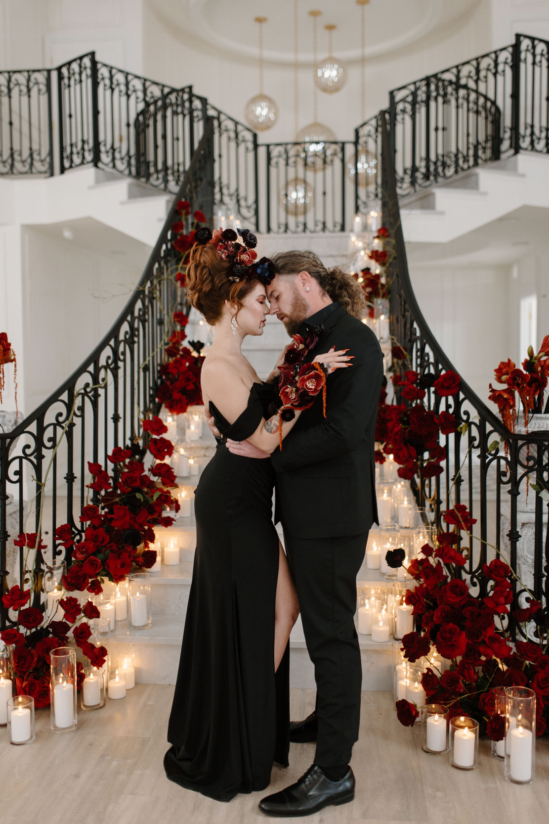 Dracula and his bride stand at the base of the stunning, dramatic staircase at Archway Manor. They are facing each other, moments away from a kiss.