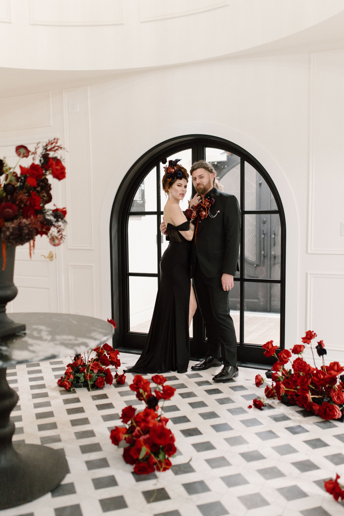 Dracula and his bride stand closely, with her hand on his chest in the gorgeous foyer at Archway Manor, with beautiful red arrangements on the stunning tiled floor.