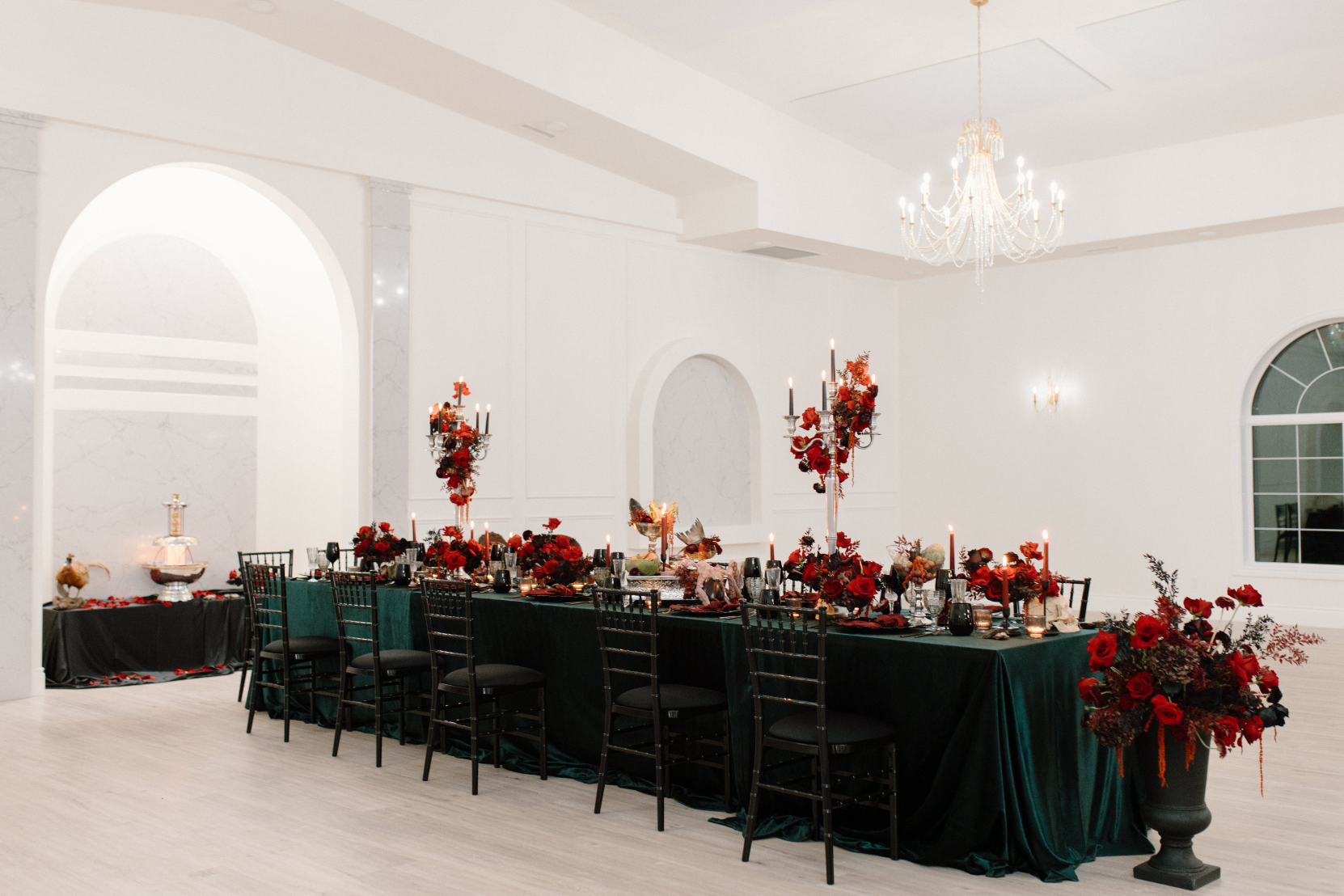 Inside Archway Manor's reception space, a long, rectangular table is beautifully styled for Dracula's deathly dinner party! Dark emerald tablecloth, black chiavari chairs, candelabras, ornate black plates, flatware and glasses covered in lavish red centrepieces, fruit and some creepy, slimy fishy things.