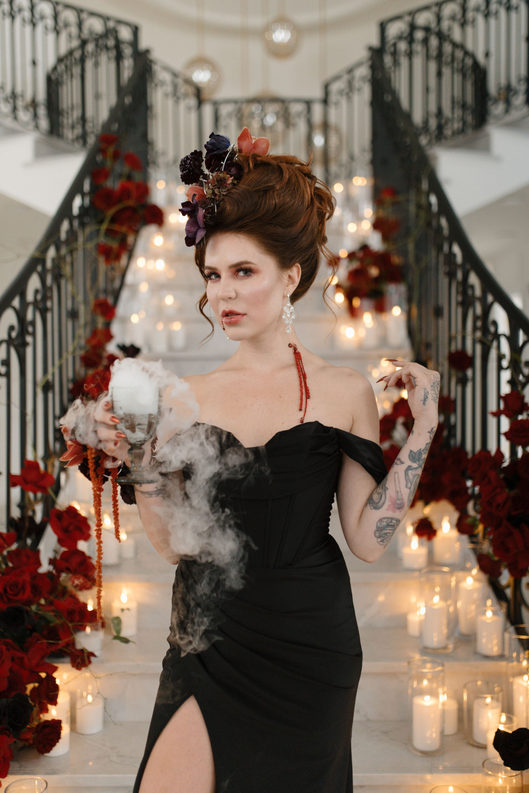 Dracula's bride standing at the staircase with a stunning updo and floral hairpiece, holding a glass overflowing with smoke.