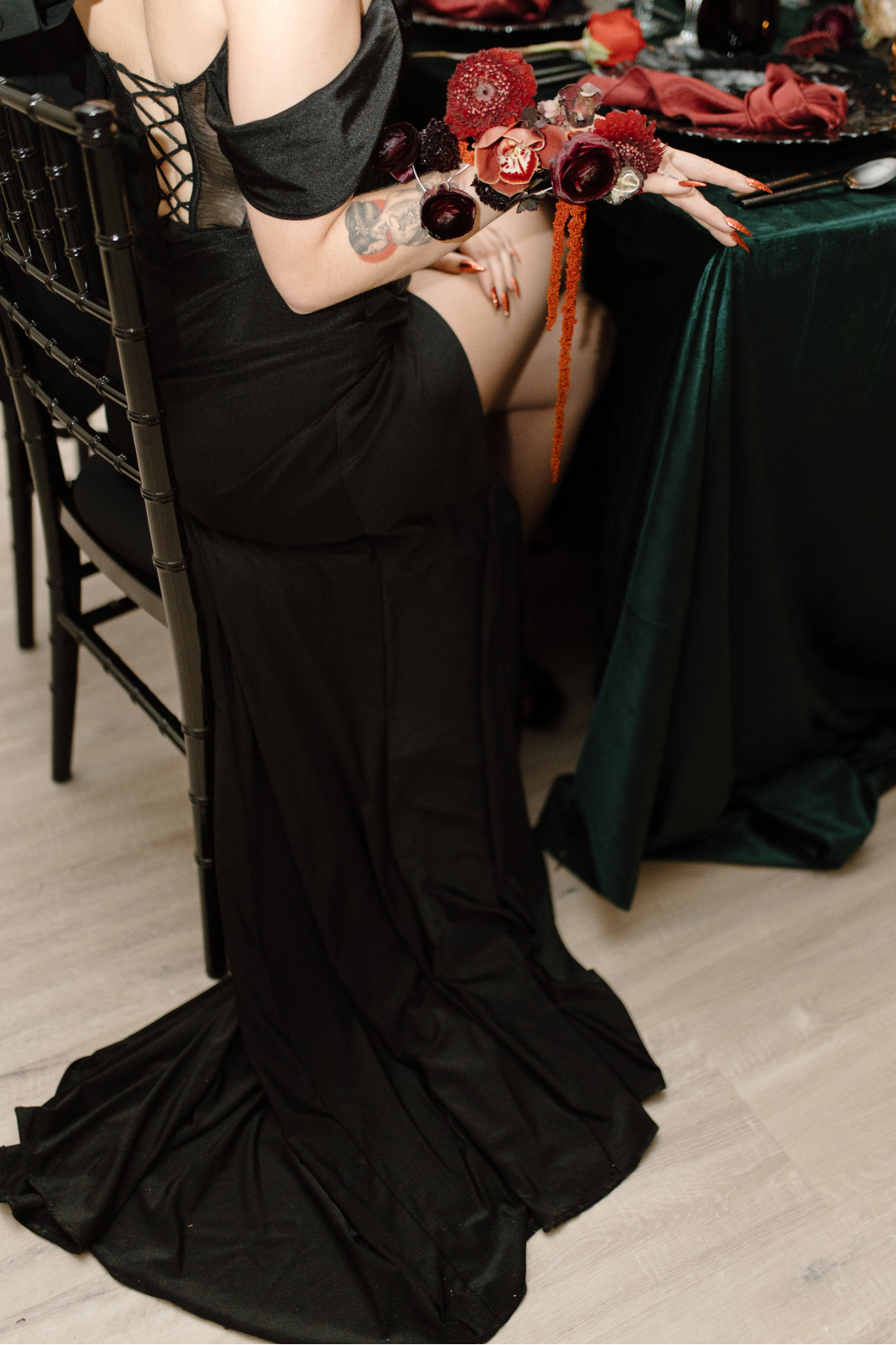 Dracula's bride sits in a chiavari chair at the table, with her dress pooled at the floor as she holds her hand out tot he side to show her lavish wrist corsage.