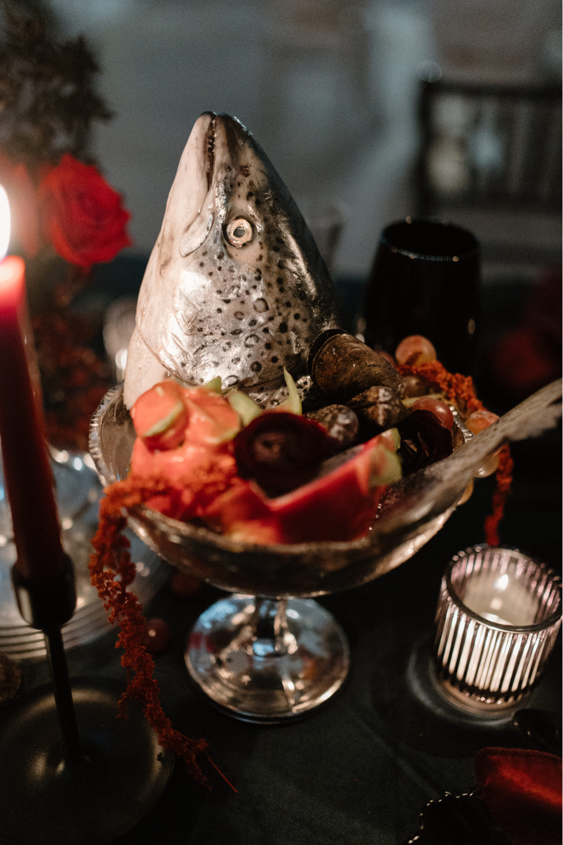 A dish holding some assorted fruit and flowers has a large fish head sticking out of it.