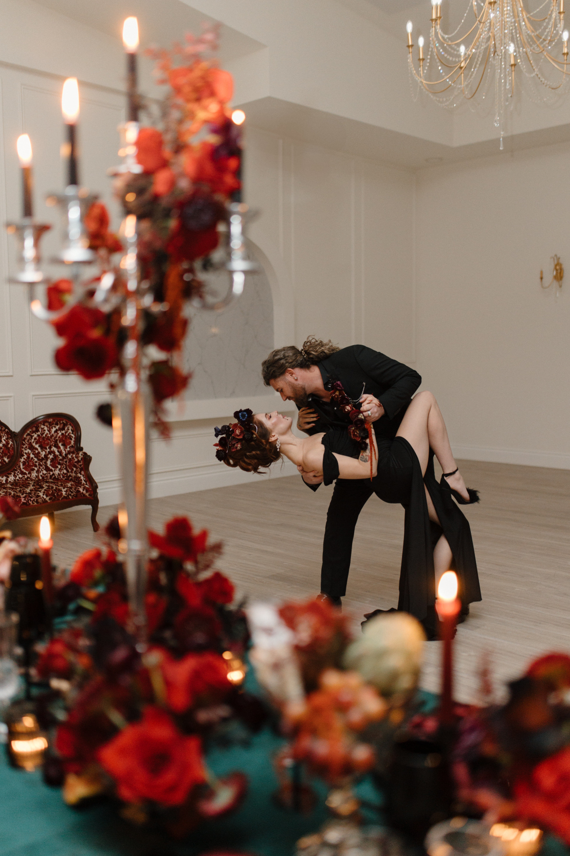 With a chandelier and floral arrangements gentle blurred, Dracula and his bride are dancing in focus behind the table, he has her dipped and her knee pulled up to his side.