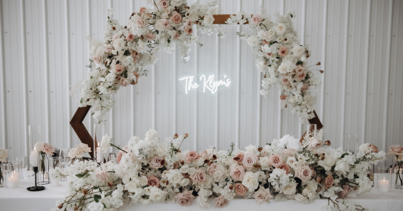 A beautiful, full arrangement of flowers in white and blush sits on the head table, behind a hexagonal wood arch, with white and blush florals with a neon sign hanging in the center that reads "The Klym's".