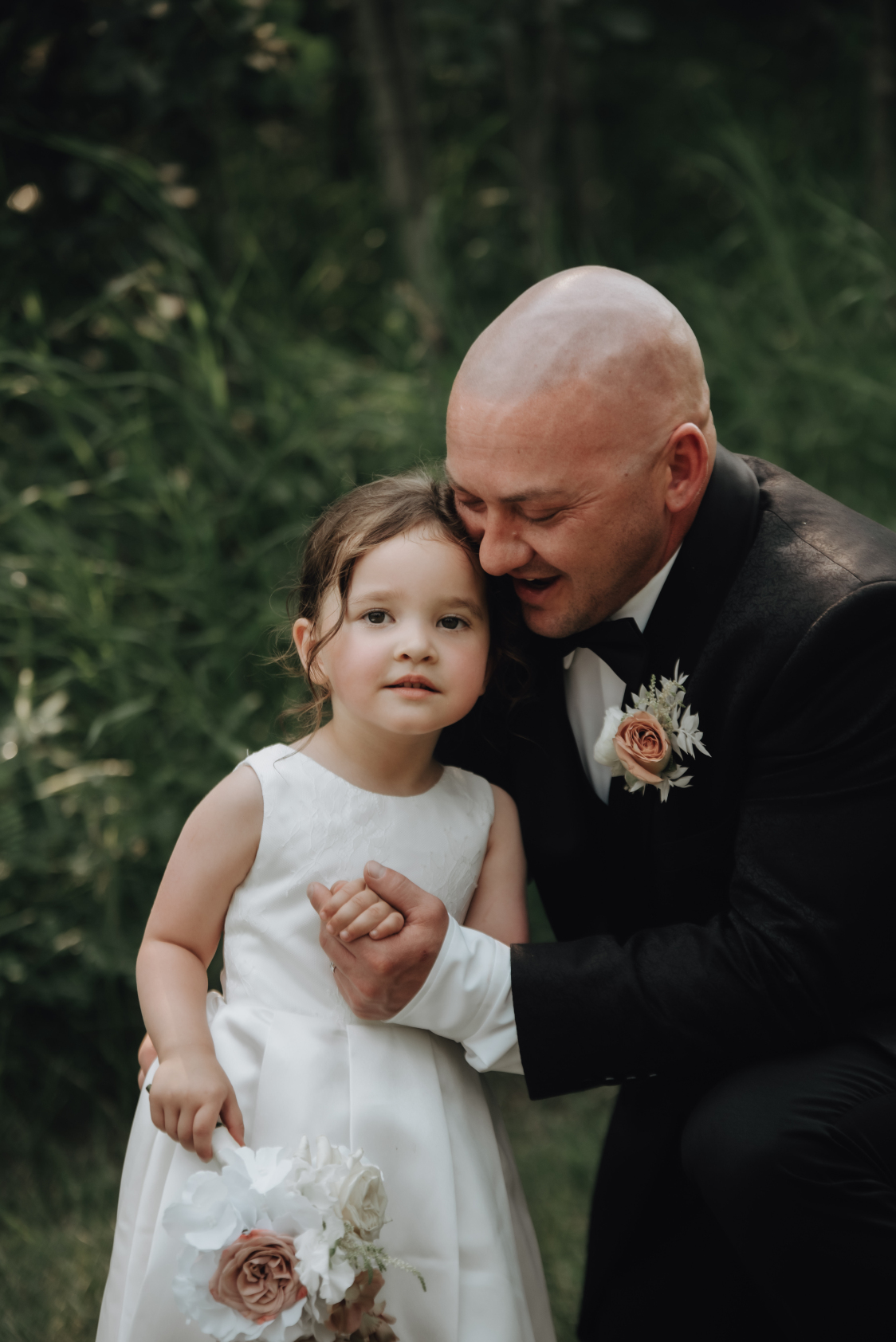 Chris snuggles up with their daughter, who is the flower girl, holding her hand and talking in her ear while she smiles at the camera