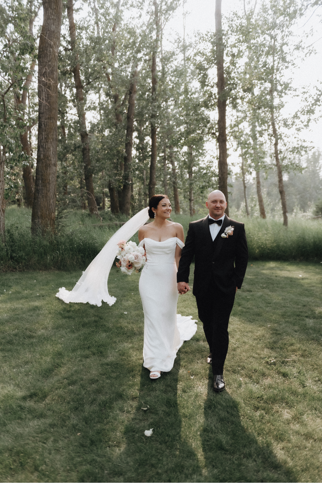 Kristen and Chris walking hand in hand in a forest like setting, her veil blowing in a breeze behind her.