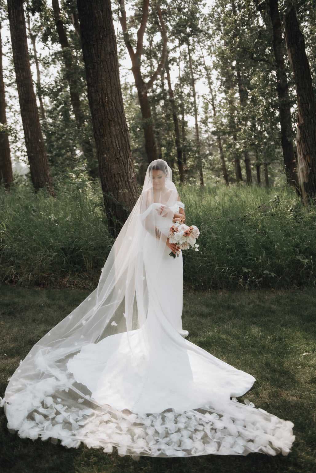Kristen stands with a treed area behind her, her dress elegantly pooled around her feet, her stunning veil covering her face and also pooled around her showing the gorgeous organza petal details