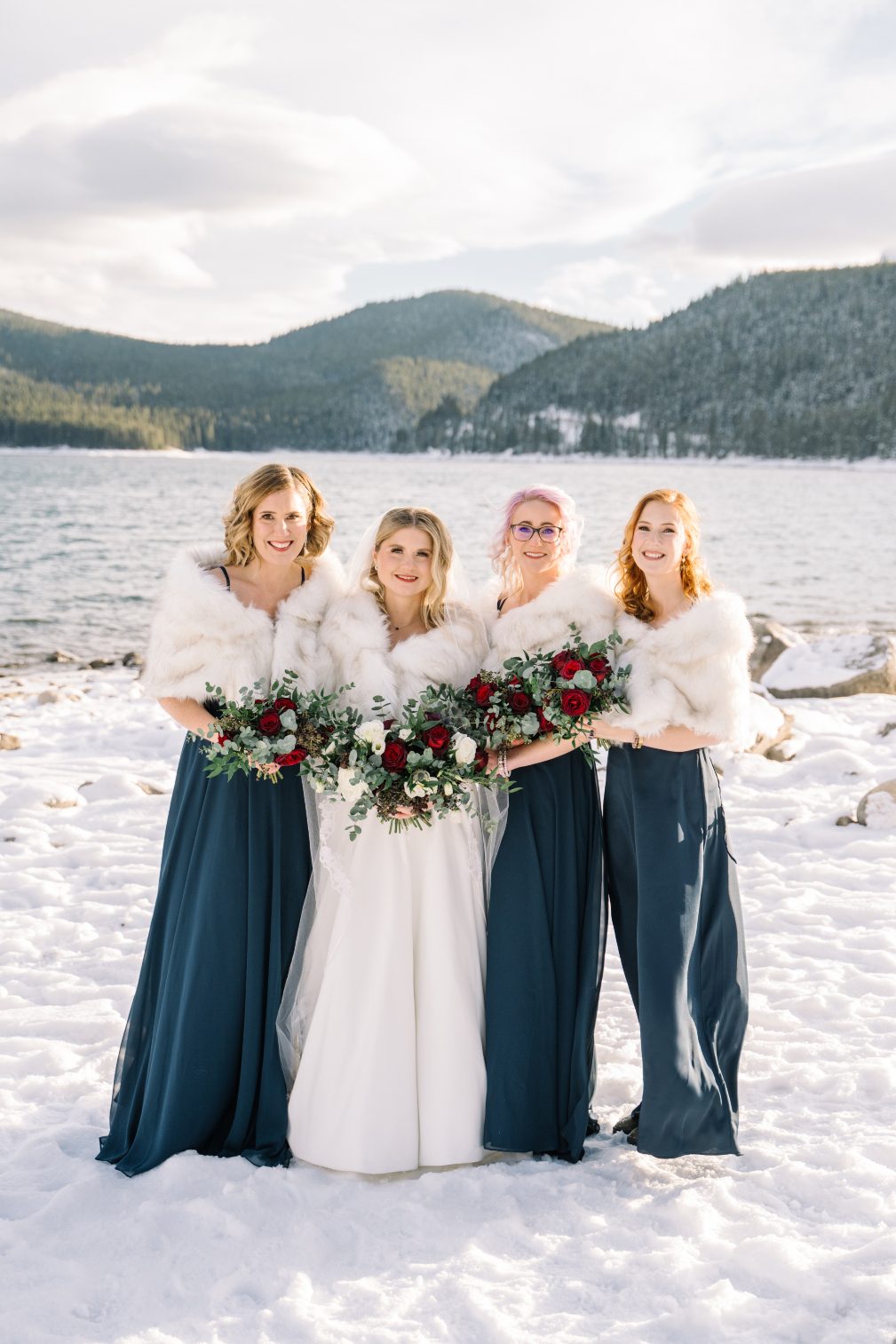 Bride and Bridesmaids Courtney and her bridesmaids, standing outside with a mountain in lake and mountains in the distance. They are draped in fur stoles and holding beautiful bouquets of burgundy and ivory.