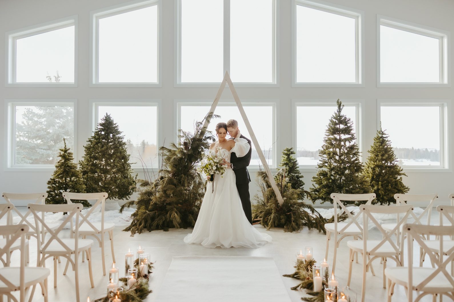 The model groom stands behind the model bride at the end of the aisle. In one hand she holds her bouquet, her other hand reaches up to touch the grooms face, which is turned towards her.