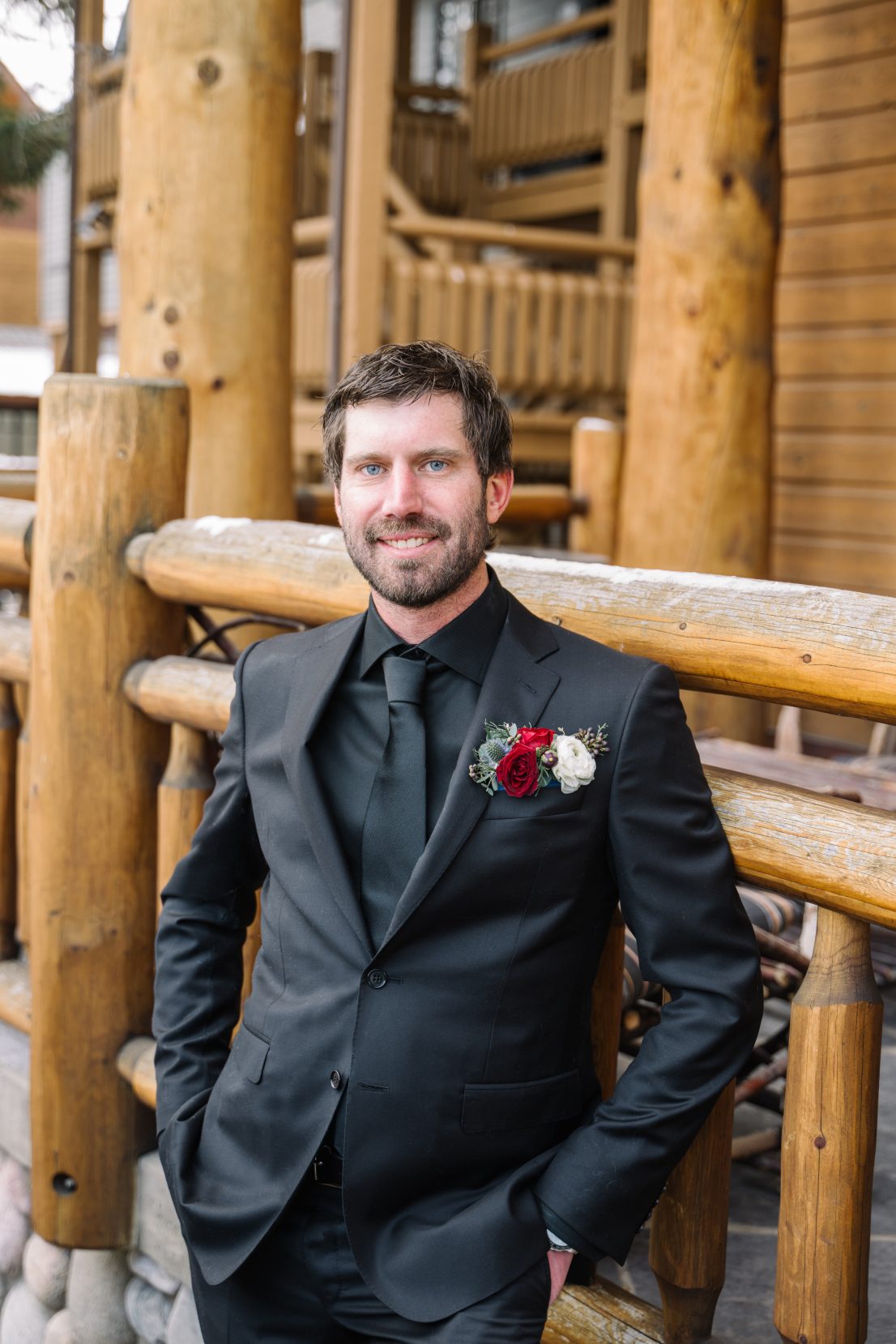 Calyx Groom, Adam Adam stands solo in front of the lodge, looking at the camera and smiling in his smart black suit and floral pocket boutonniere.
