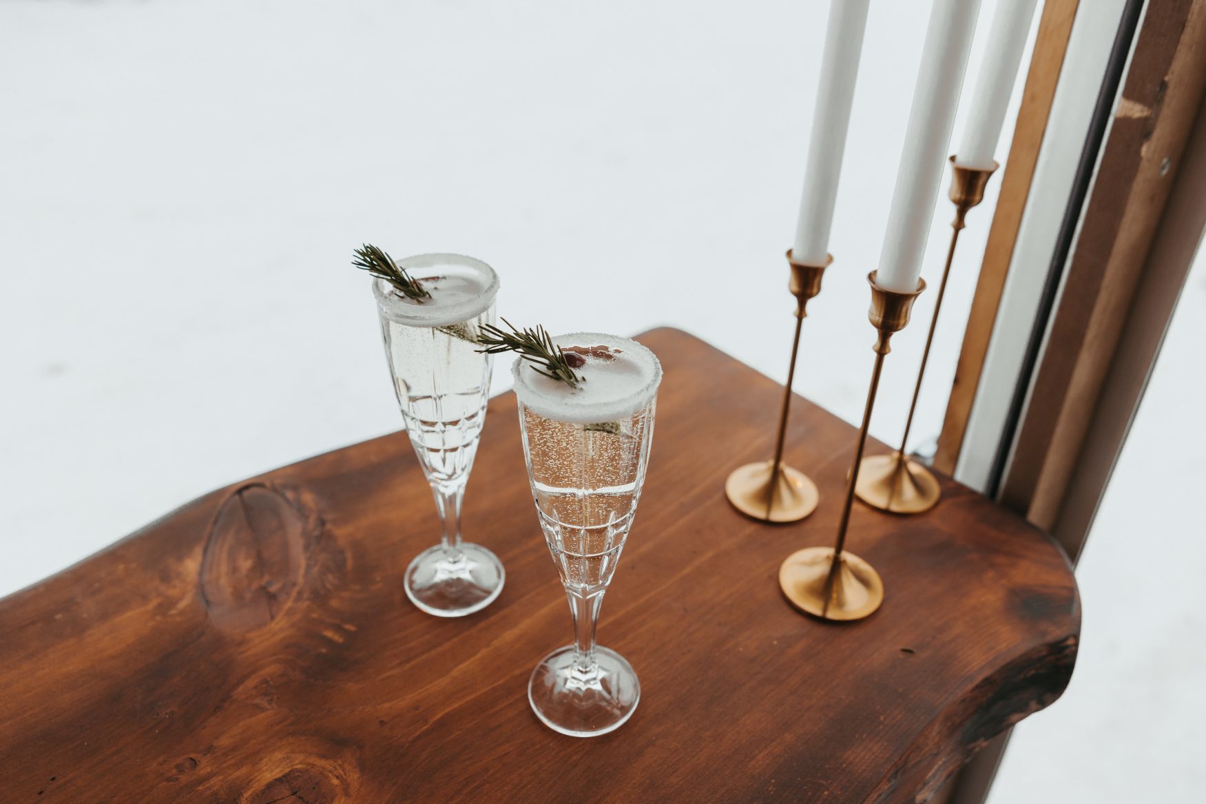 Two glasses of champagne garnished with a couple cranberries and a sprig of rosemary sit on top of a wood table with next to white tapered candles.