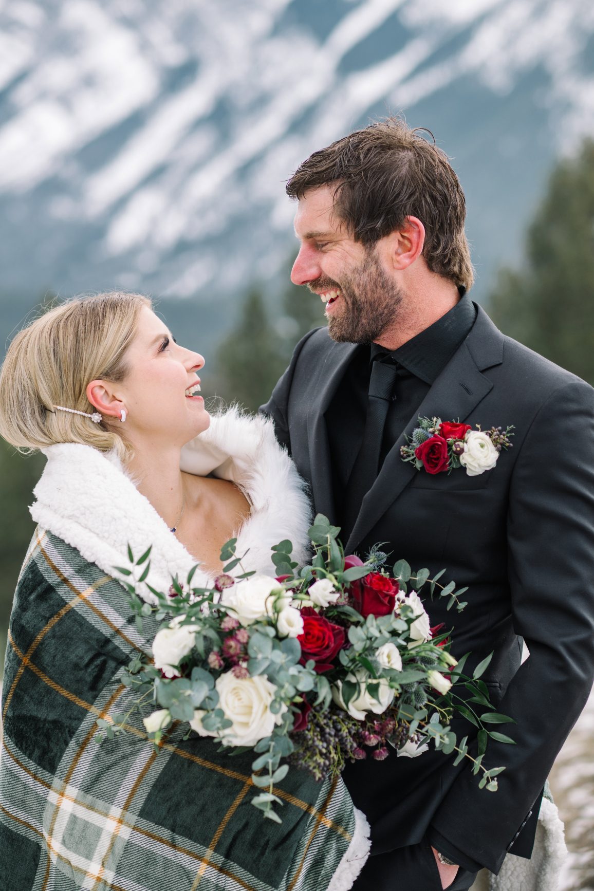 Calgary Wedding and Portrait Photographer Courtney has a plaid wrap covering her shoulders as she holds her stunning bouquet. Her and Adam are looking at each other and smiling.
