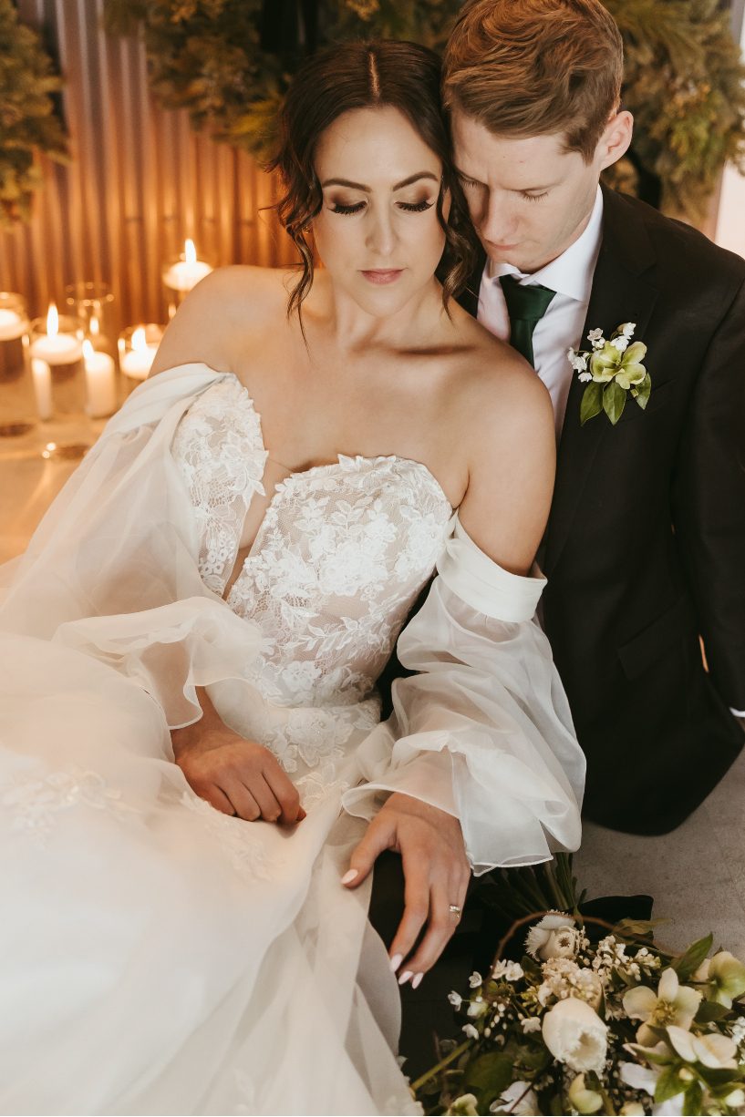 The couple sit together on the floor, the bride nestled into his chest with her bouquet by her side with lit candles in the background.