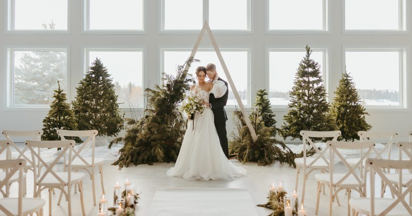 Model bride and groom embrace each other at the end of the aisle in front of evergreen trees, a triangle arch, and huge windows.