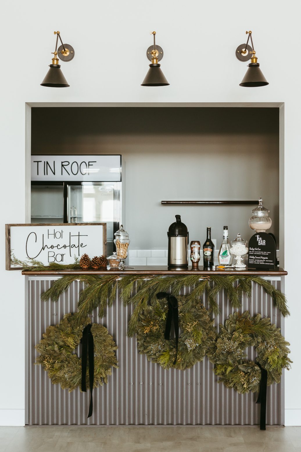 A cute little bar set up, decorated with three hanging wreaths and some branches of pine, has a sign reading "hot chocolate bar" and has assorted hot chocolate goodies displayed on the counter.