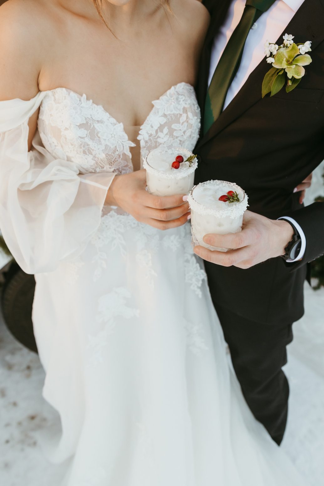 The happy couple standing closely, each handing their own mug of delicious looking cocktails with decorative rims and garnished with a couple cranberries and a sprig of rosemary.