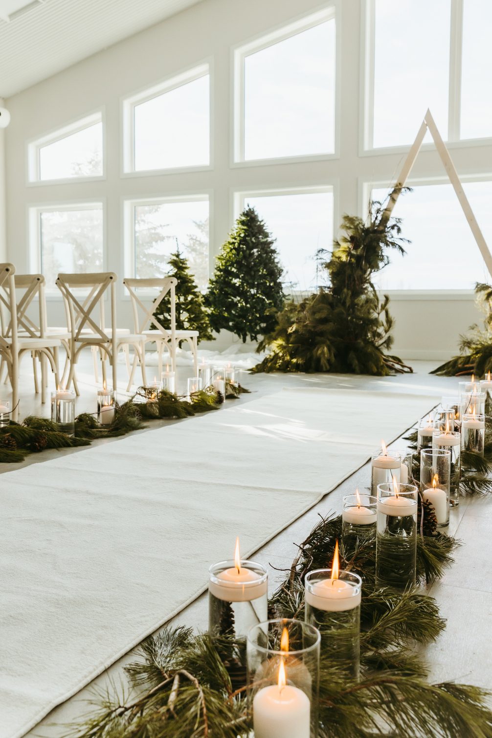 The aisle decorated with gorgeous pine, cylinder vases with candles and the and of aisle decorated with a triangle arch adorned with assorted winter greens.