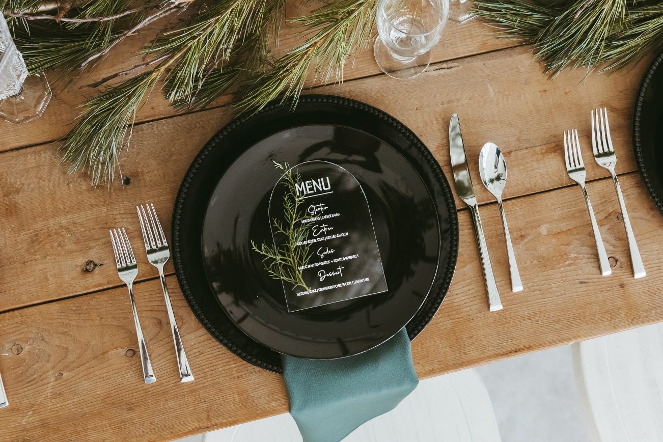 Gorgeous wood table displaying a guest place setting with black plates, a sheer pine coloured napkin, a small sprig of juniper and a clear acrylic sheet with the menu etched onto the top.