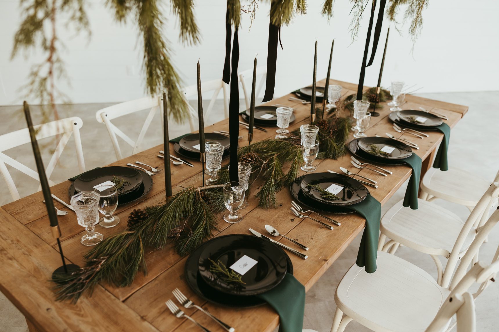 The view of the center of the table underneath the centrepiece, with gorgeous dark coloured tapered candles, pine branches, black plates, sheer pine coloured napkins and name cards on each plate.