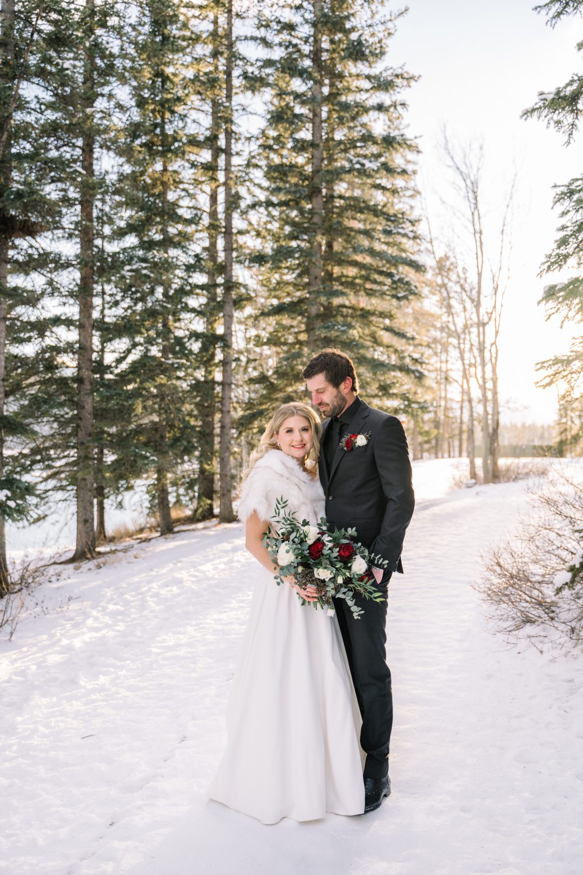 Winter Couple Standing in a treed area, with the soft glow of sunlight on them, Courtney and Adam are snuggled up close as he gazes lovingly at her.