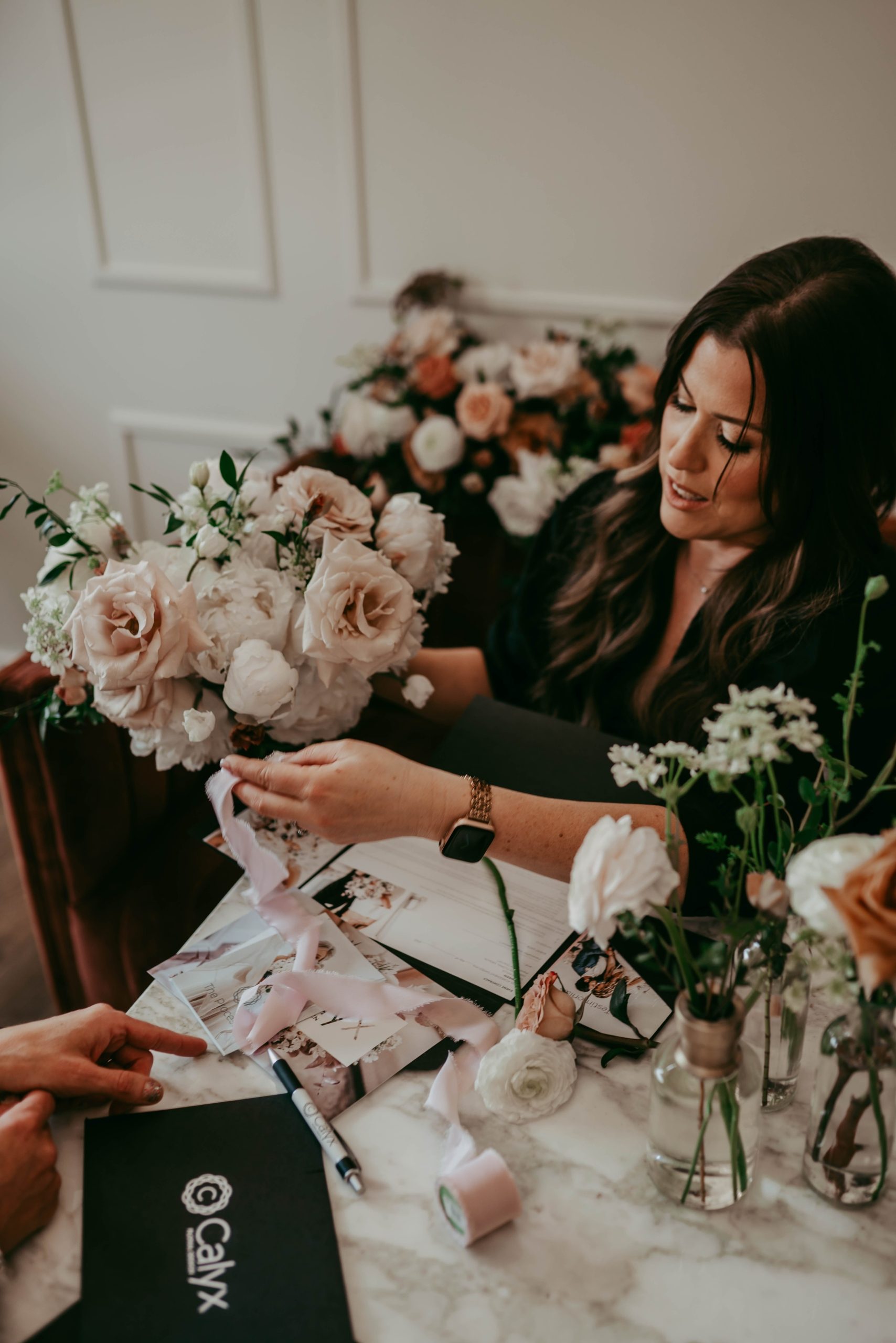 Travis planning out a wedding, holding a bridal bouquet and coordinating silk ribbon, with planning materials scattered on the desk.