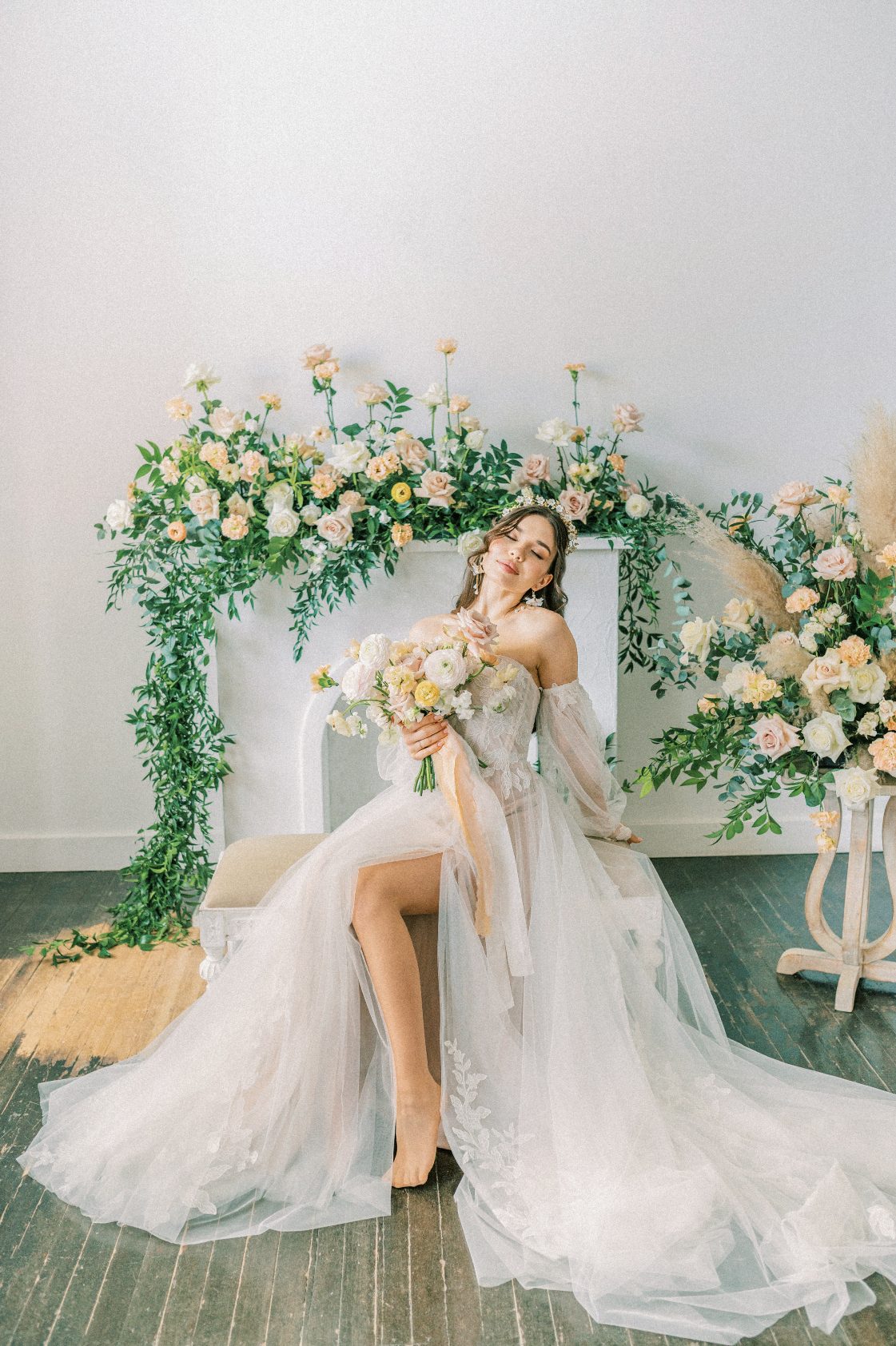 A model bride sits on a bench with her dress splayed out beautifully. she holds a stunning Calyx bouquet in pastel tones, with stunning matching arrangements displayed behind her.