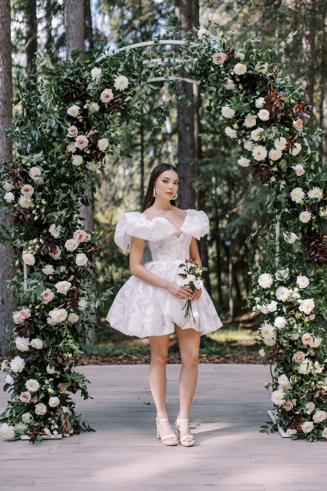 A model stands under the outdoor arch with a super cute short bridal gown with stunning ruffle accent acting as sleeves.